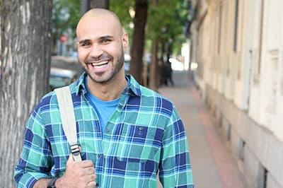 smiling man with backpack standing on a sidewalk next to a building